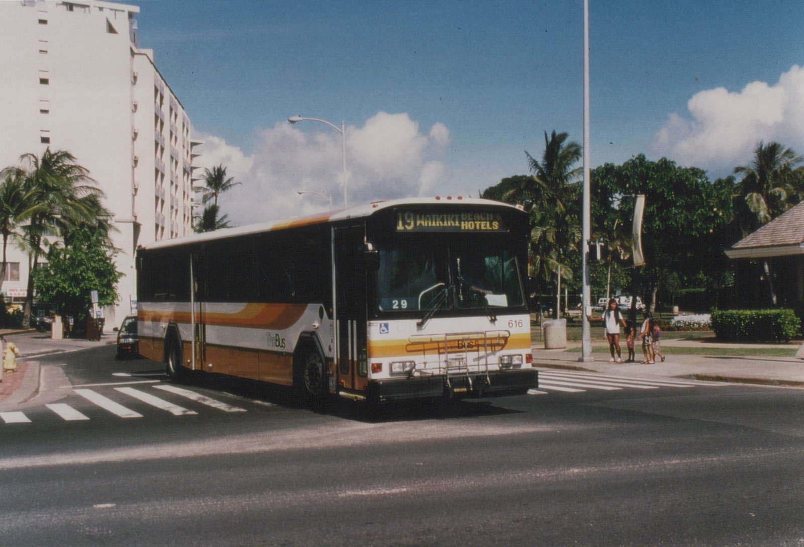 A white and orange TheBus, number 616, displays route 19 to Waikiki Beach and Hotels as it drives down a palm-lined street with pedestrians crossing nearby.