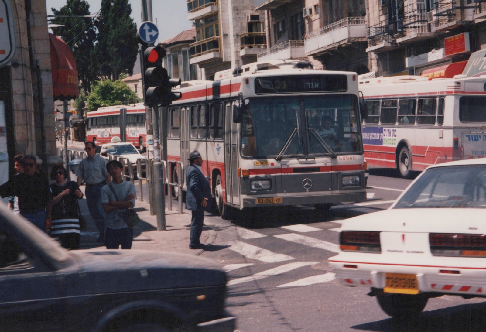A white and red Egged bus is stopped at a red light on a busy urban street in Israel, with people on the sidewalk and other vehicles around it.