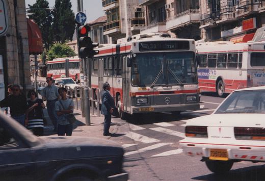 A white and red Egged bus is stopped at a red light on a busy urban street in Israel, with people on the sidewalk and other vehicles around it.