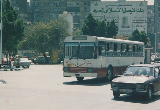 A white and red motor bus with the fleet number 4873 is driving on a city street, with a brown sedan in the foreground and a large Arabic billboard on a building in the background.
