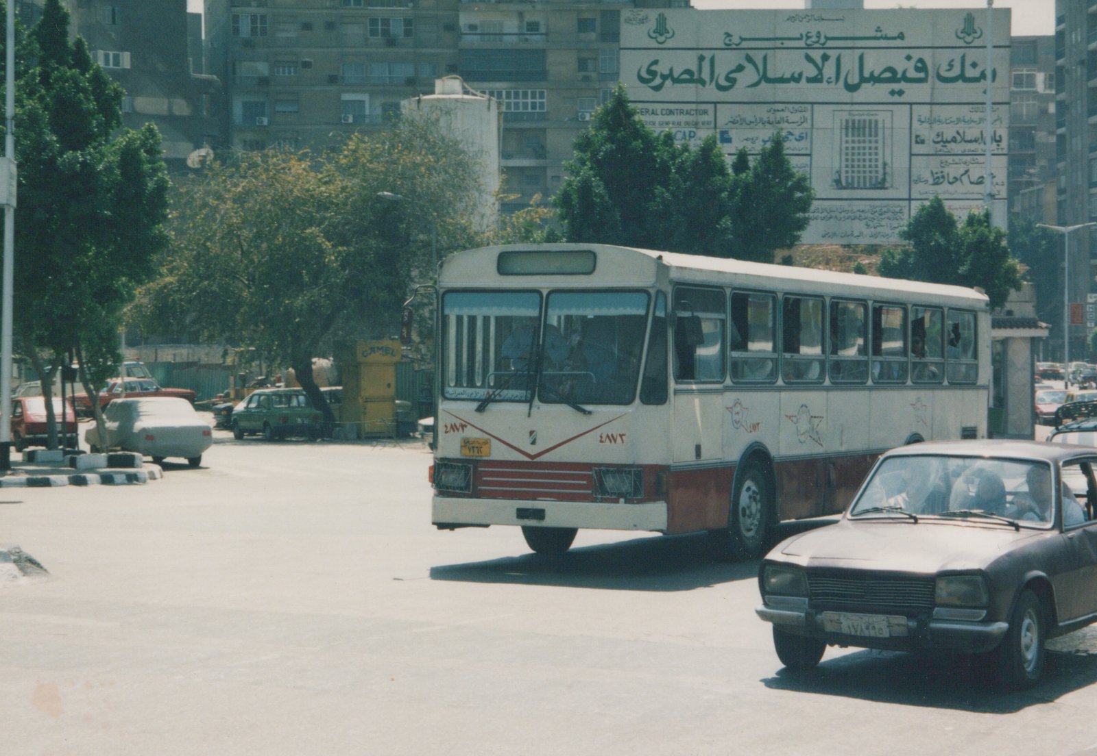 A white and red motor bus with the fleet number 4873 is driving on a city street, with a brown sedan in the foreground and a large Arabic billboard on a building in the background.