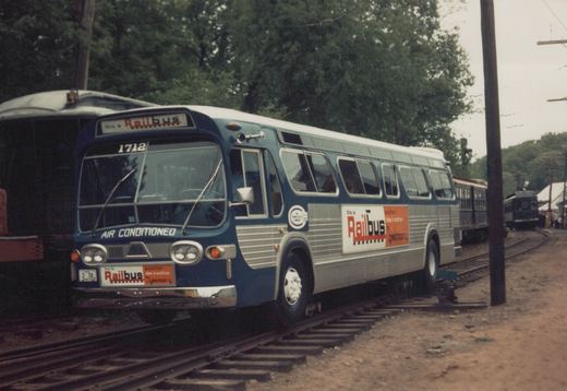 A blue and silver motor bus labeled 'Railbus' is positioned on railroad tracks, with old interurban cars visible behind it.