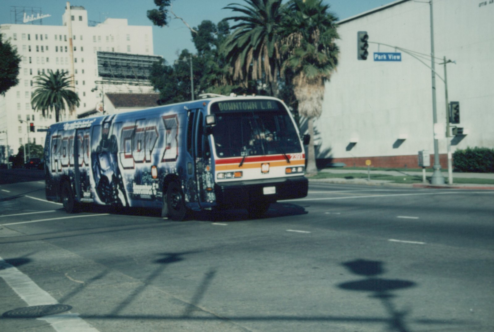 A blue and white bus with a 'RoboCop 3' ad on its side drives down a street lined with palm trees, with a 'Park View' street sign visible.
