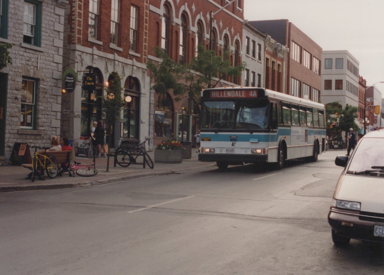 A white and blue Kingston Transit bus, number 9297, driving down a city street lined with brick buildings and pedestrians.