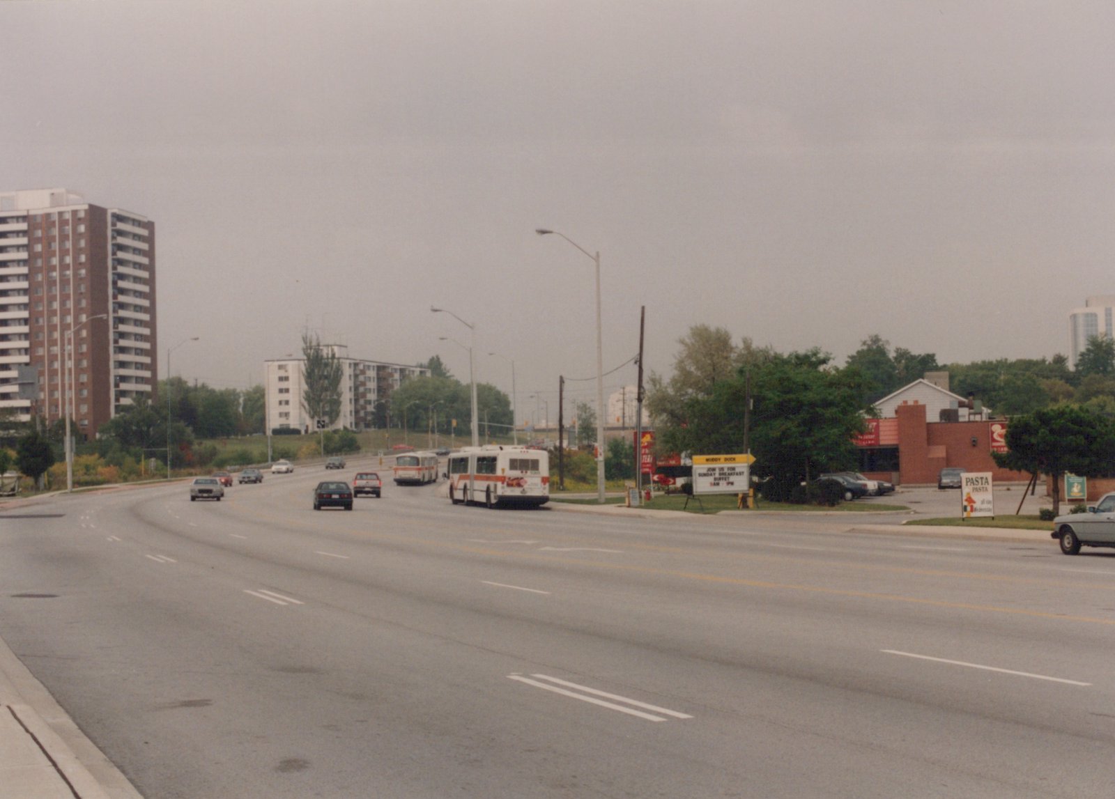 A wide, overcast street with two white and orange buses, several cars, apartment buildings, and restaurant signs, including one for Muddy Duck and another for Pasta Pasta.