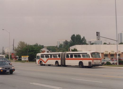 An articulated white and orange bus with the number 5016 is driving down a multi-lane road in a suburban area with overcast skies, past a restaurant and other cars.