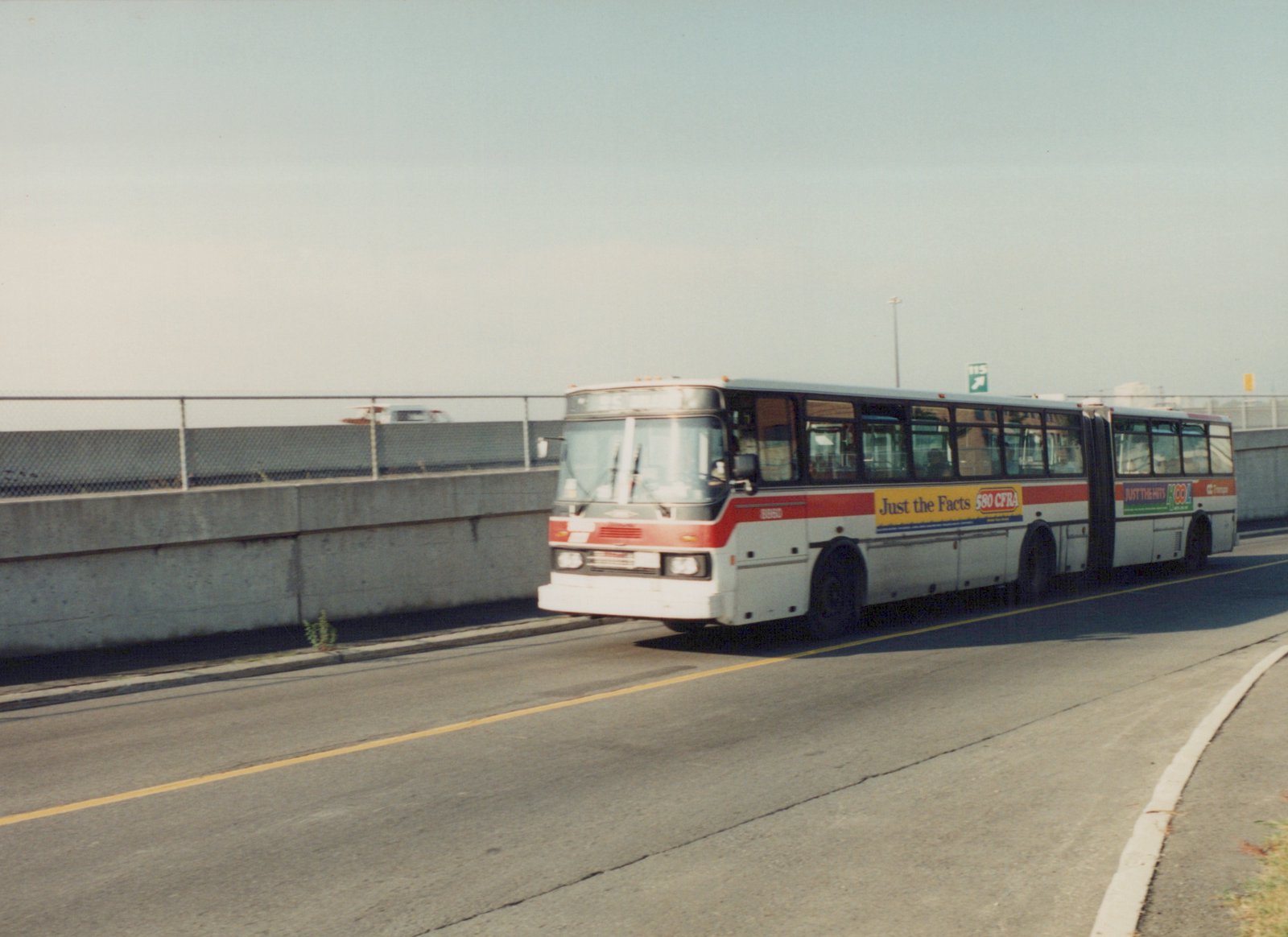 A white and red articulated bus with the number 8950 on its side drives on a highway with a concrete barrier and a chain-link fence in the background.