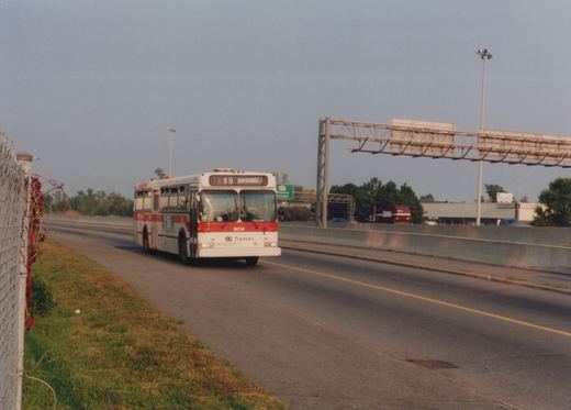 A white and red OC Transpo bus drives on a highway median with a green highway sign in the background.