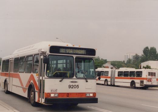 A white and orange Mississauga Transit bus, number 9205, labeled '81 VIA DUNDAS' on its front sign, drives down a street with another articulated bus in the background under an overcast sky.