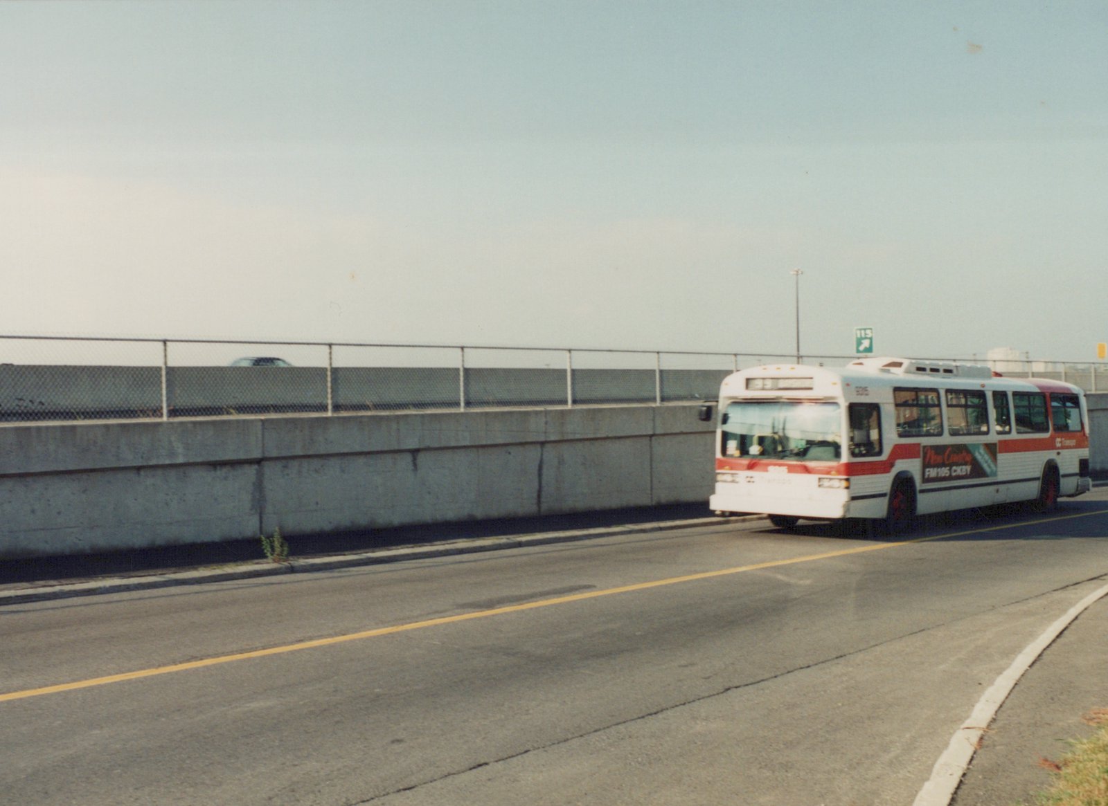 A white and red OC Transpo motor bus, number 935, drives on a road beside a concrete wall under a clear sky.