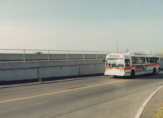 A white and red OC Transpo motor bus, number 935, drives on a road beside a concrete wall under a clear sky.