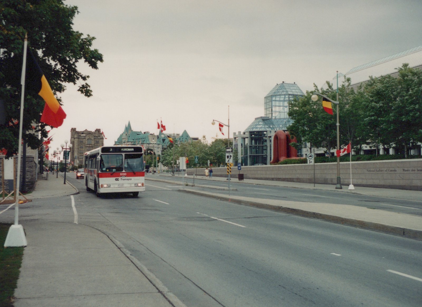 A white and red OC Transpo city bus drives down a tree-lined street in Ottawa, with Canadian and Belgian flags and the National Gallery building in the background.