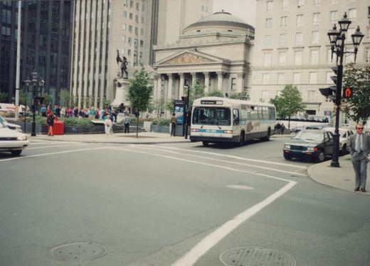 A white and blue city bus with the number 55-061 is turning at a city intersection in front of a large classical-style building, with pedestrians and other cars nearby.