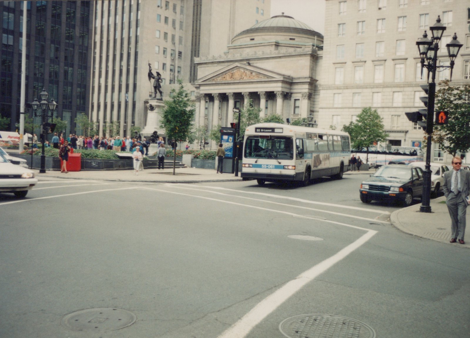A white and blue city bus with the number 55-061 is turning at a city intersection in front of a large classical-style building, with pedestrians and other cars nearby.