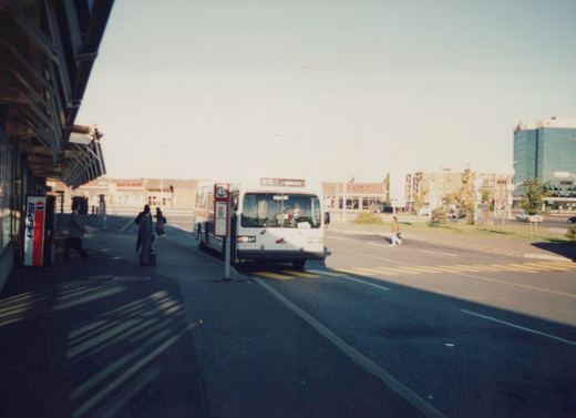 A white bus with red and blue stripes is stopped at a bus stop on a sunny day, with several people nearby and a Pepsi vending machine visible on the left side of the image.