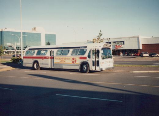 A side-front view of a white bus with a red stripe and a sail logo, number 7-794, driving on a street with a Nautilus Plus store and modern buildings in the background.