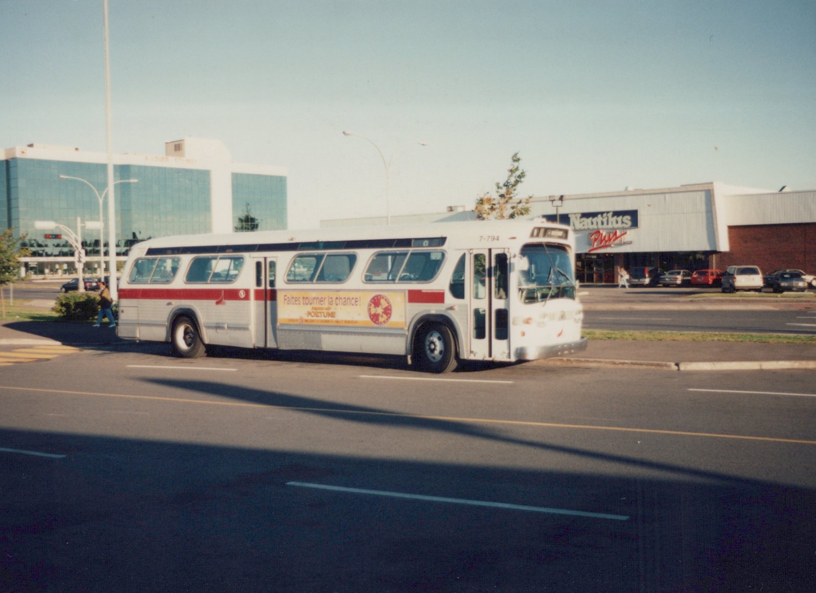 A side-front view of a white bus with a red stripe and a sail logo, number 7-794, driving on a street with a Nautilus Plus store and modern buildings in the background.