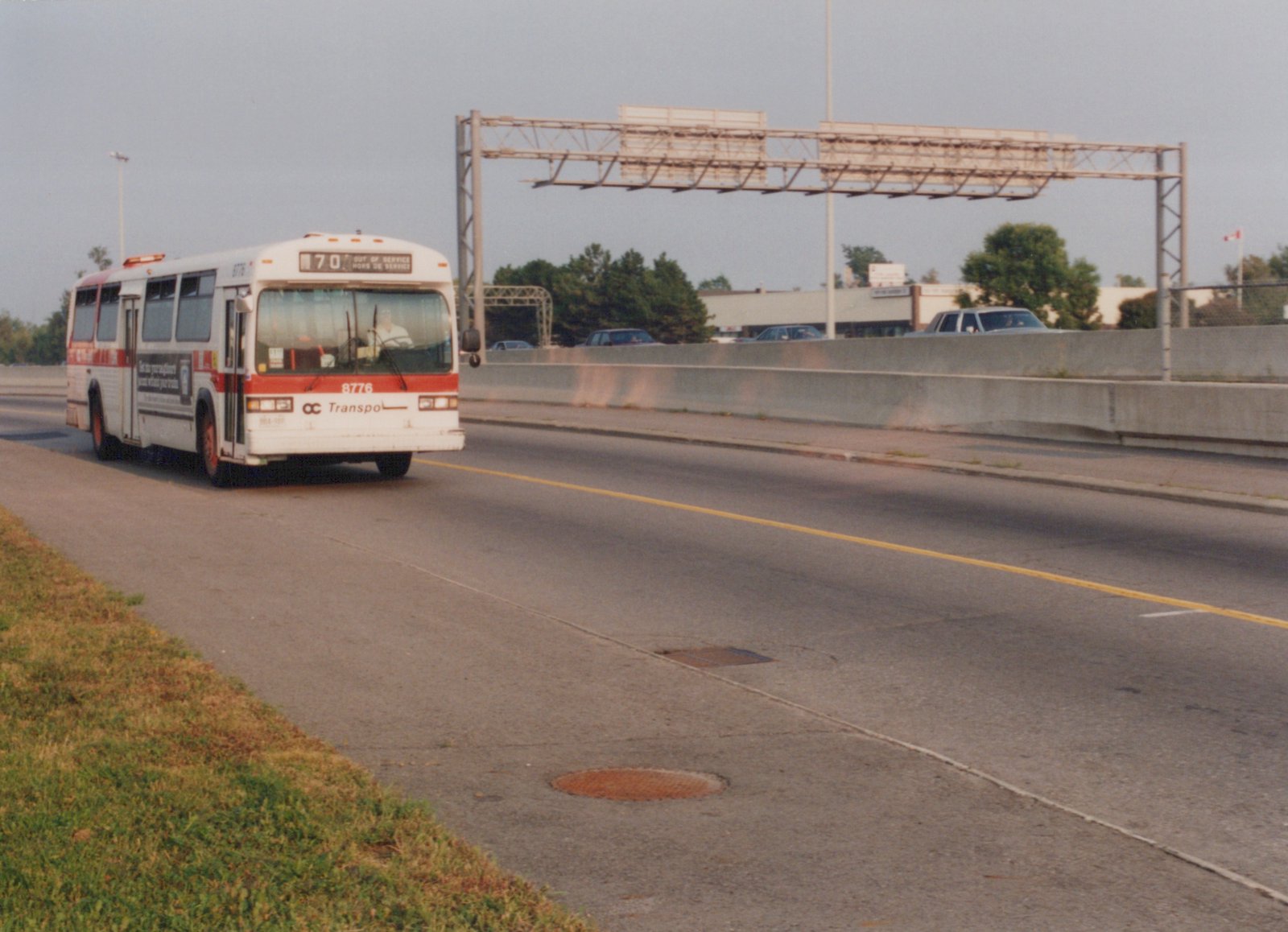A white and red OC Transpo bus, number 8776, labeled 'Out of Service,' drives on a highway median with a concrete barrier and green trees in the background.