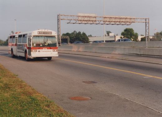 A white and red OC Transpo bus, number 8776, labeled 'Out of Service,' drives on a highway median with a concrete barrier and green trees in the background.