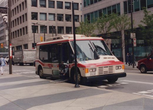 A white and red motor bus with WHEEL lettering on the side is stopped on a city street with its ramp down, surrounded by pedestrians and buildings.