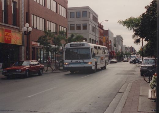 A white MCI bus with 'Barriefield 6' on the destination sign drives down a city street lined with brick buildings, parked cars, and a pedestrian on the sidewalk.