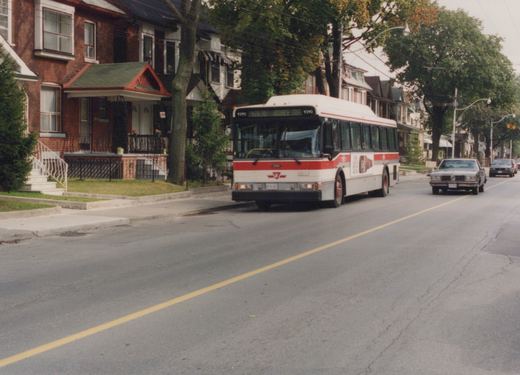 A white and red Toronto Transit Commission bus, number 9390, is driving down a tree-lined residential street with houses on the left and a few cars behind it.