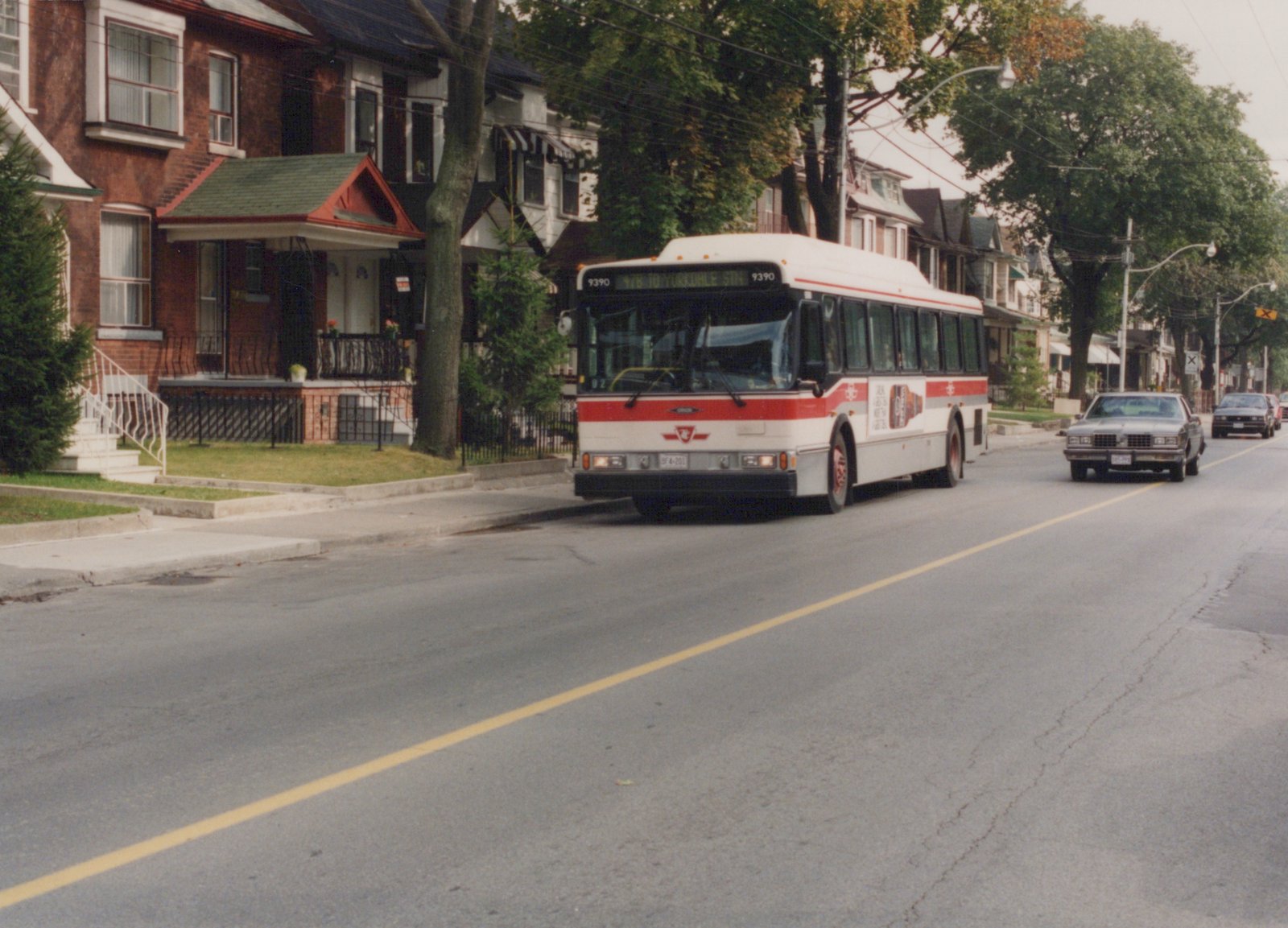 A white and red Toronto Transit Commission bus, number 9390, is driving down a tree-lined residential street with houses on the left and a few cars behind it.