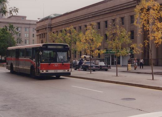 A red and grey Toronto Transit Commission bus numbered 6054 is driving on a city street lined with trees showing autumn colors, with a large stone building and pedestrians in the background.
