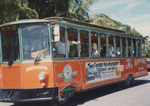 A side view of an orange and green Old Town Trolley tour bus, number 82, driving on a sunny street lined with palm trees in Key West, Florida, with several passengers visible through the windows.