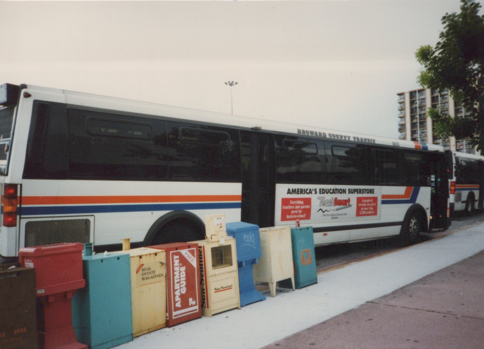 A white, orange, and blue Broward County Transit bus, number 9665, is parked on a city street next to a row of colorful newspaper and magazine dispensers, with another bus visible behind it and a high-rise building in the background.