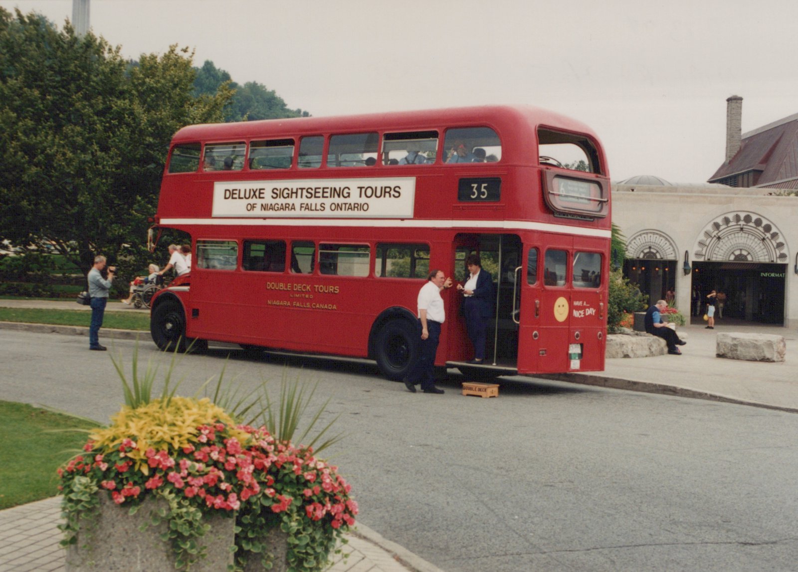 A red double-decker tour bus with 'Deluxe Sightseeing Tours of Niagara Falls Ontario' on its side is parked on a street, with passengers boarding and people nearby.