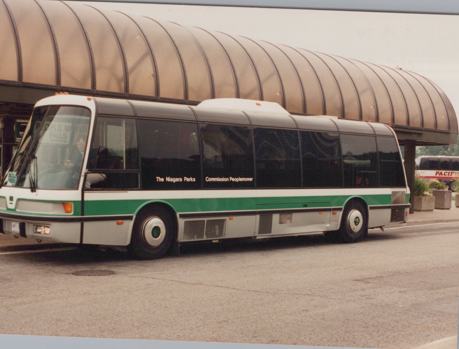A white and green motor bus with 'The Niagara Parks Commission Peoplemover' lettering is parked on a street under a curved canopy. A driver is visible inside. A second bus with 'PACIFIC' on its side is in the background.