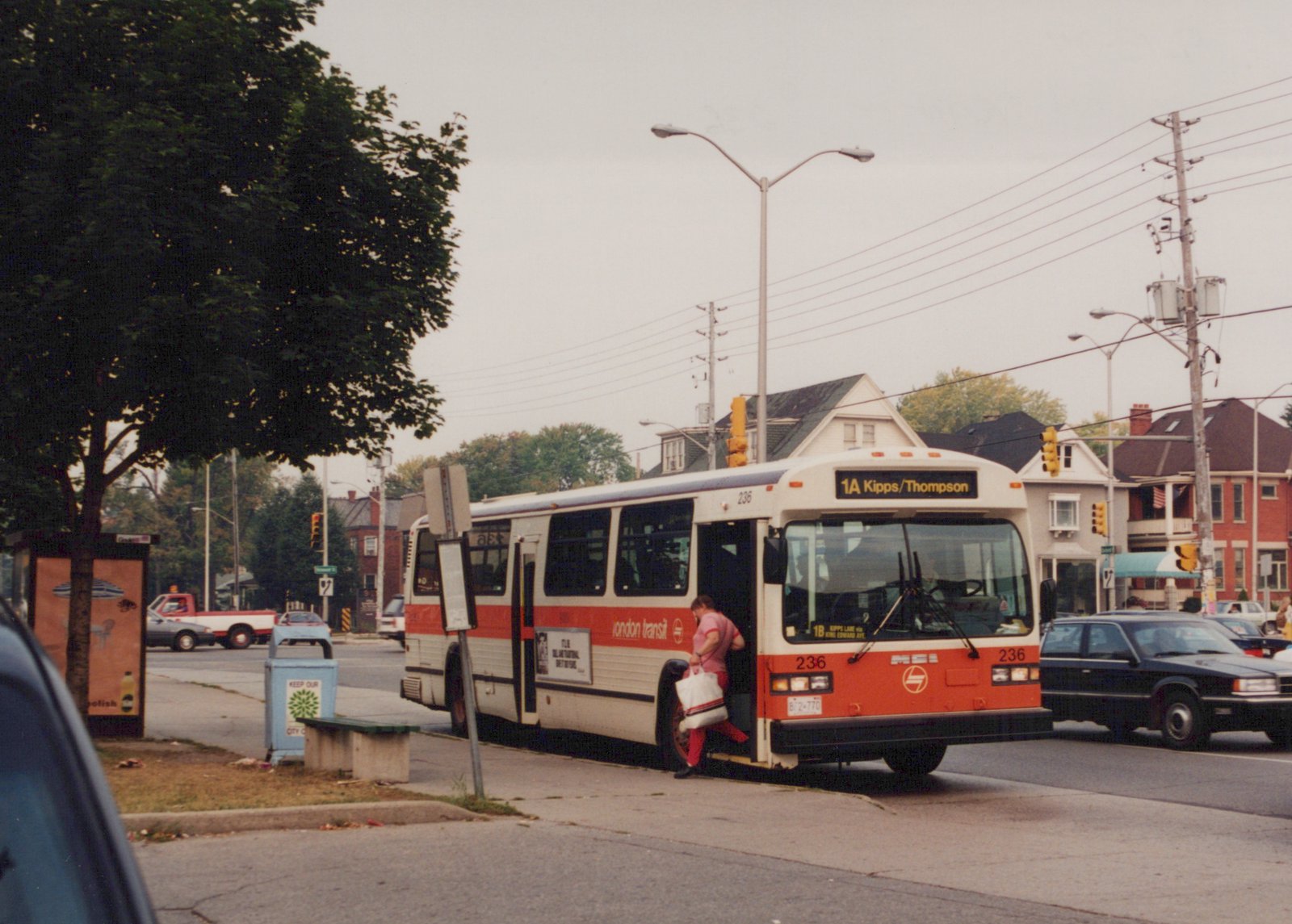 A white and orange London Transit bus, number 236, is stopped on a city street with a passenger stepping aboard; the route sign reads '1A Kipps/Thompson' and the scene is set against a backdrop of residential houses and traffic lights.