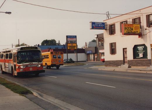 A red and white city bus numbered 145 drives down a paved street with a yellow school bus in the background and storefronts for Bondi's Pizza and Shaker Auto Body on the right.