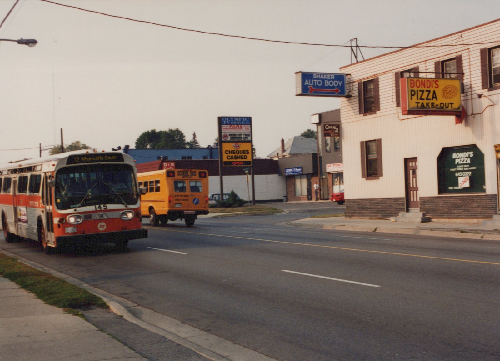 A red and white city bus numbered 145 drives down a paved street with a yellow school bus in the background and storefronts for Bondi's Pizza and Shaker Auto Body on the right.