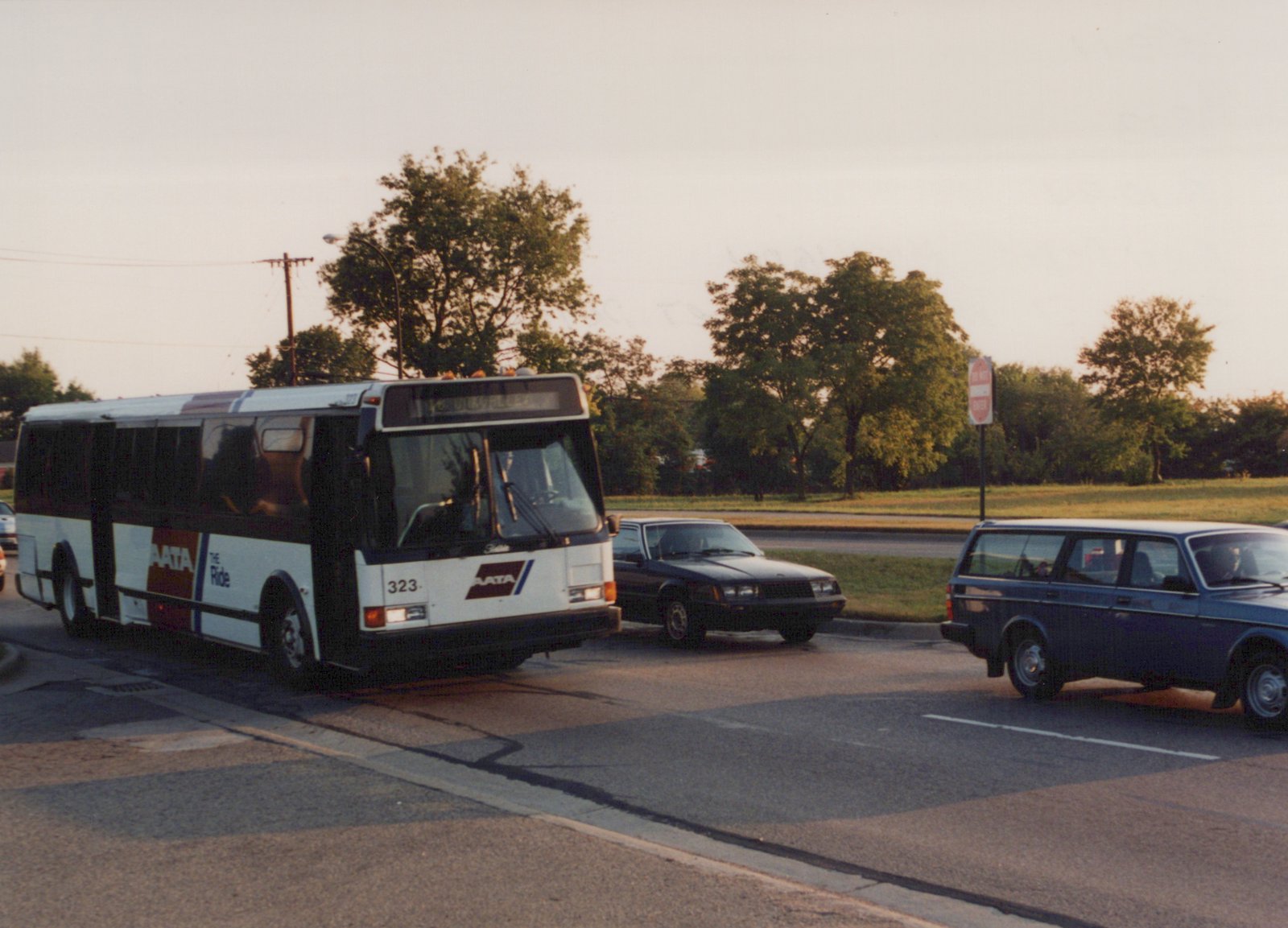 A white and maroon AATA bus with the number 323 is turning a corner on a street, with cars and a "Do Not Enter" sign in the background.