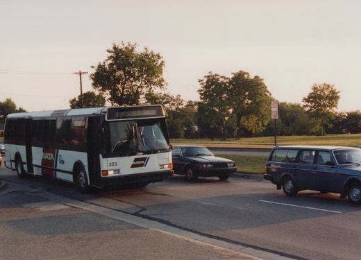 A white and maroon AATA bus with the number 323 is turning a corner on a street, with cars and a "Do Not Enter" sign in the background.