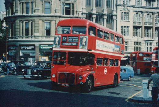 A red double-decker bus with route 12 to Harlesden, marked RM491, drives on a London street with classic black cabs and storefronts in the background.