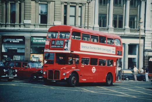 A classic red double-decker bus in London, with passengers boarding and people on the street, set against a historic building facade.