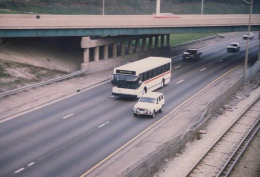 A white CTA bus with an orange stripe and a sign reading 'WHO'S IN FIRST?' drives on a multi-lane highway. Other cars are on the road and on an overpass above. Train tracks run parallel to the highway on the right.