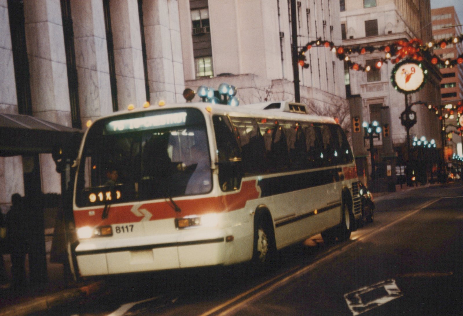 A white and red CTA bus with the number 8117 on the front, displaying route 76 to Chestnut on its sign, driving on a city street at dusk with Christmas lights on the buildings.