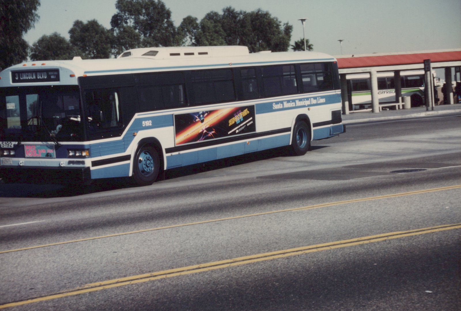 A white and blue Santa Monica bus, number 5192, is shown from the side on a sunny day, with a 'Star Trek' ad on its side and a terminal in the background.