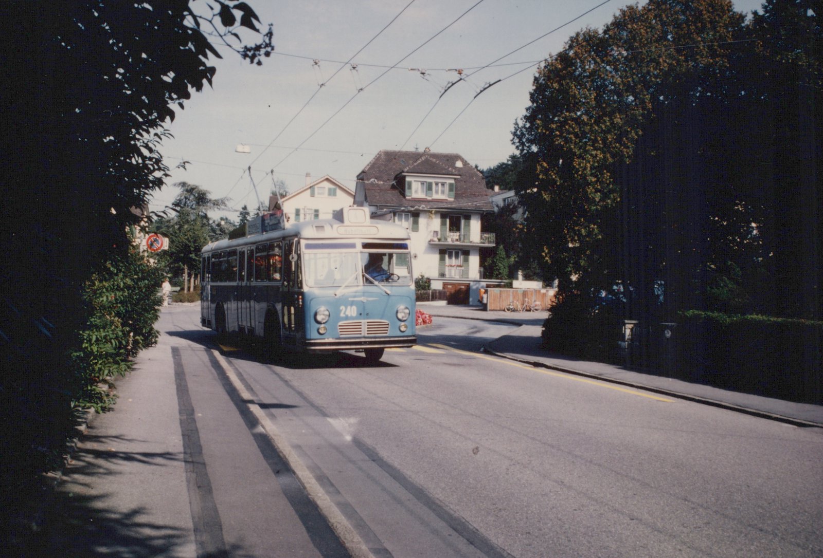 A light blue and dark blue trolleybus with the number 240 and destination 'Höhenmatt' is driving on a tree-lined street, powered by overhead wires.