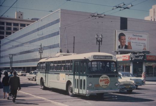 A light green and white trolleybus with number 9124 and a 'Bubble Up' ad on the front, running on a city street with overhead wires, a billboard for Calvert Reserve, and pedestrians walking nearby.