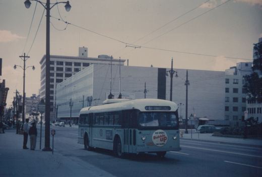 A light green and white trolleybus with the number 9025 on the front, running on a city street with overhead wires, with two people standing on the sidewalk and a billboard in the background.