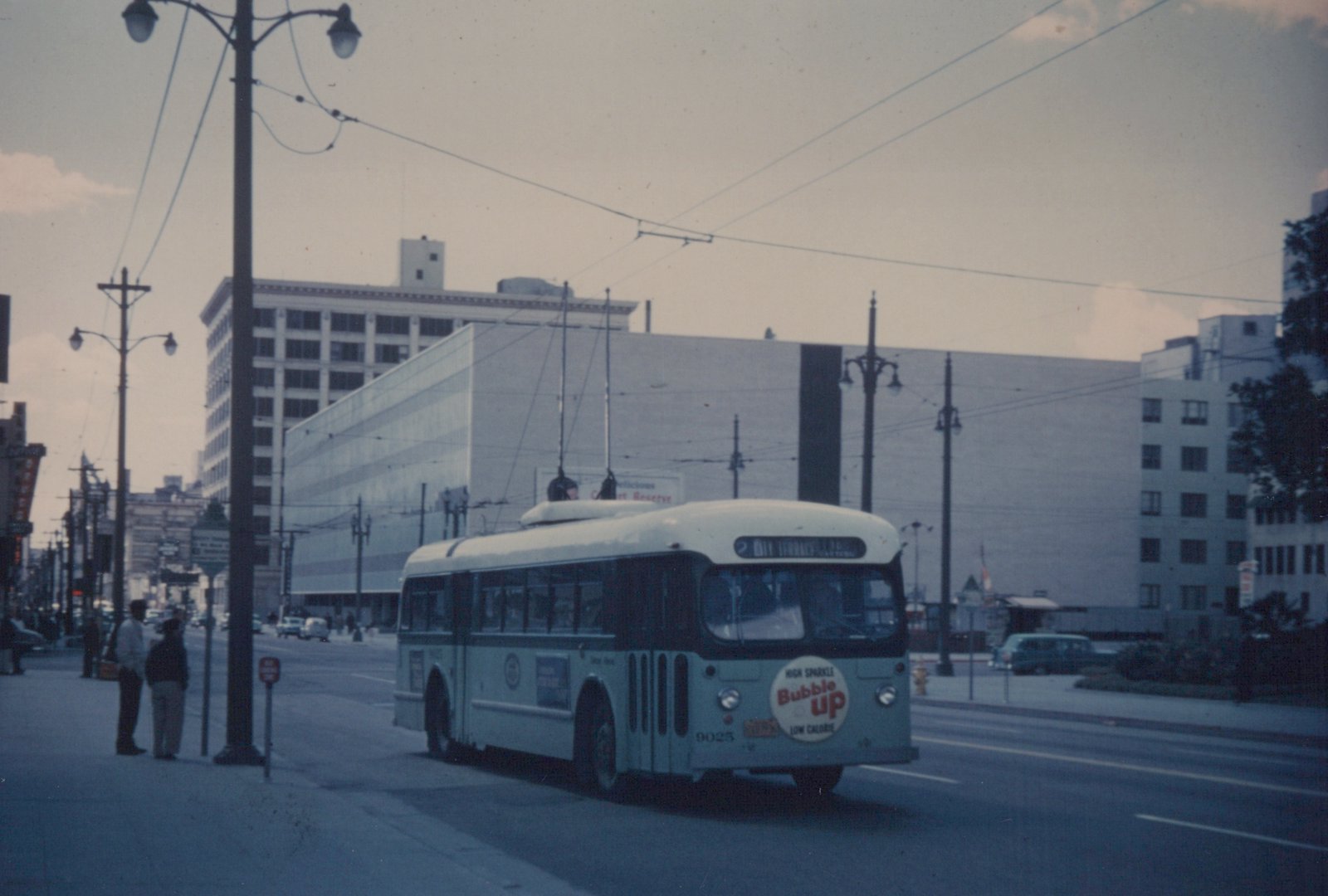 A light green and white trolleybus with the number 9025 on the front, running on a city street with overhead wires, with two people standing on the sidewalk and a billboard in the background.