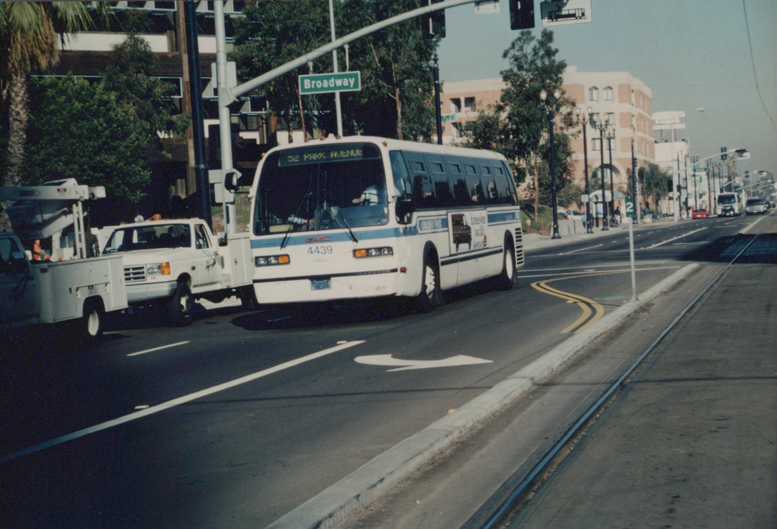 A white and blue motor bus with the number 4439 and a destination sign for 52 Park Avenue is driving on a street named Broadway, with a utility truck and trolley tracks visible.