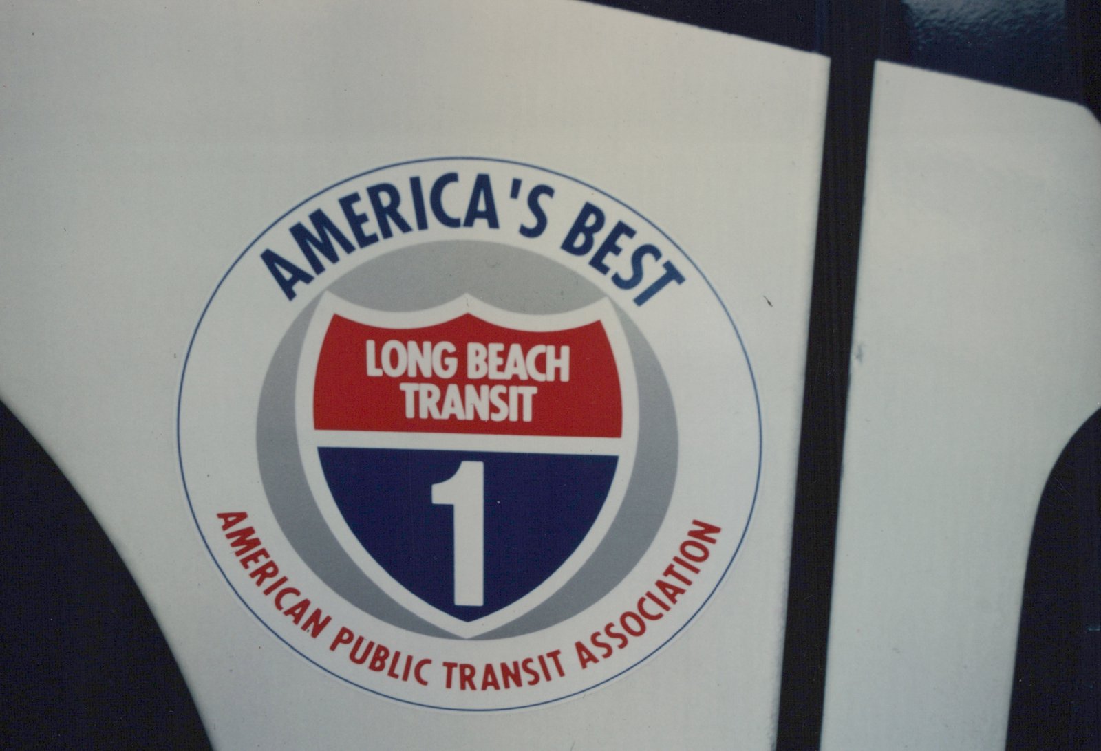 Close-up of a circular decal on a white surface. The decal has a blue border with 'AMERICA'S BEST' on top and 'AMERICAN PUBLIC TRANSIT ASSOCIATION' on the bottom. In the center is a shield with 'LONG BEACH TRANSIT' in white text on a red background and a large white number '1' on a blue background.