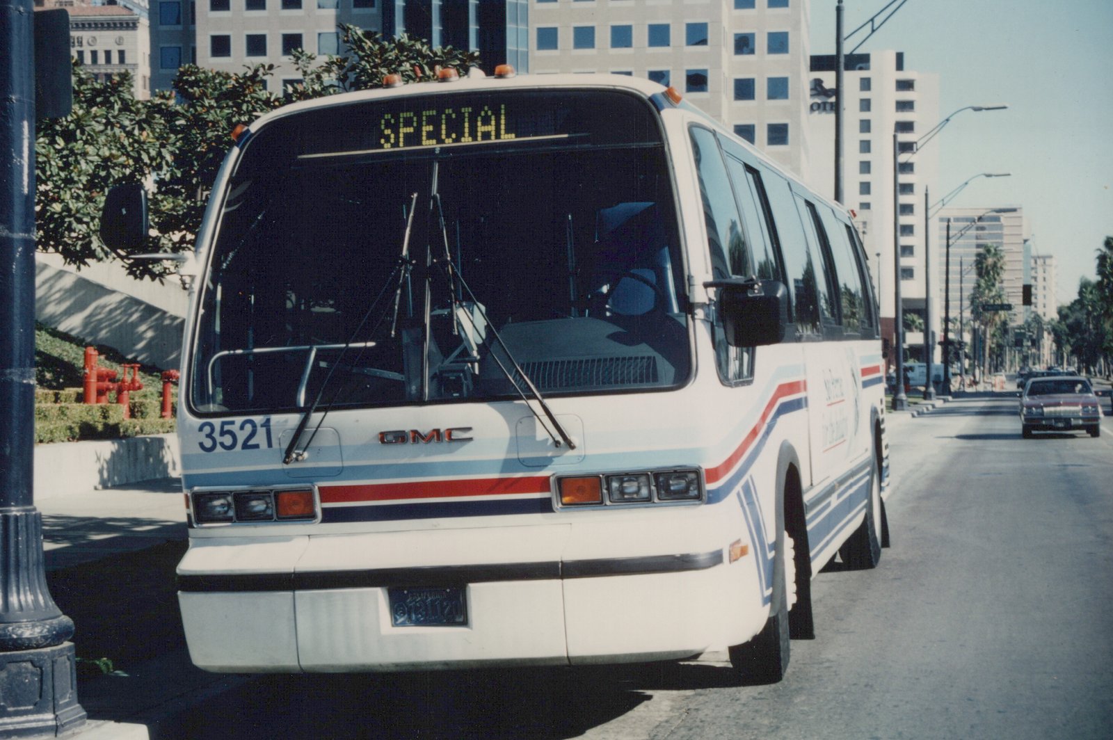Front three-quarter view of a white GMC New Look motor bus with red, white and blue stripes, showing fleet number 3521 and a 'SPECIAL' destination sign, parked on a city street with a red car and buildings in the background.