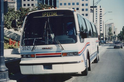 Front three-quarter view of a white GMC New Look motor bus with red, white and blue stripes, showing fleet number 3521 and a 'SPECIAL' destination sign, parked on a city street with a red car and buildings in the background.